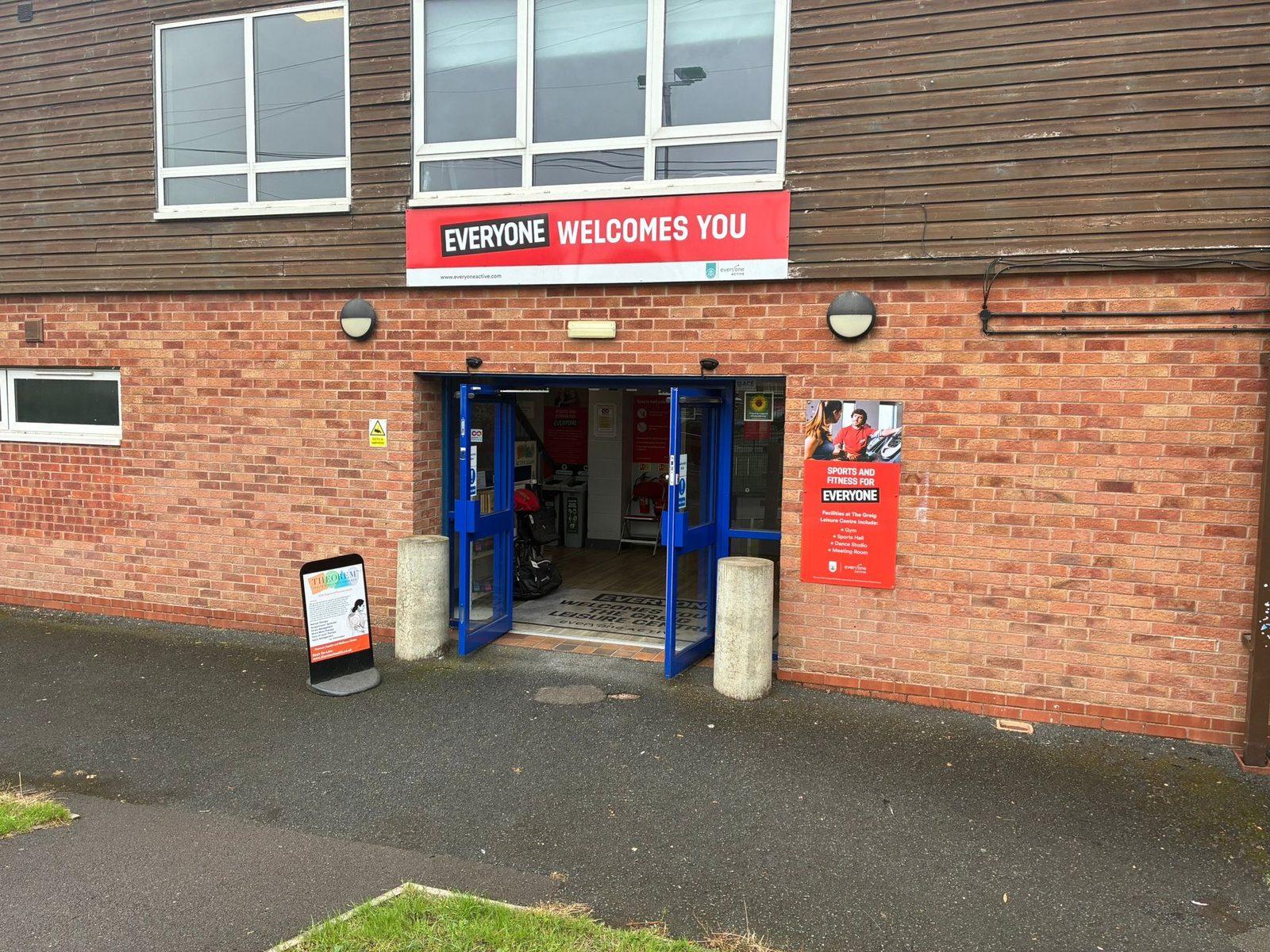 The blue double doors of the main entrance to The Greig Leisure Centre in Alcester, beneath a red Everyone Welcomes You banner